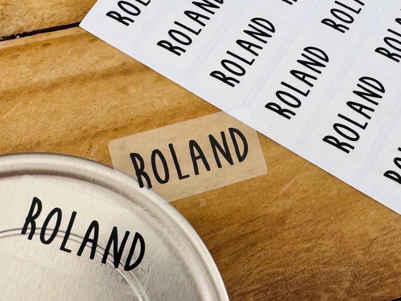 May include: A sheet of rectangular adhesive labels and a single detached label, each featuring the name ROLAND printed in a bold, black, sans-serif font. The labels are transparent, allowing the surface underneath to be visible through them.