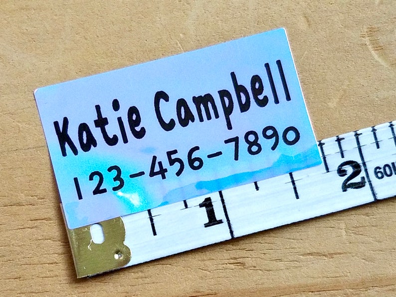 K&ouml;nnte beinhalten: Ein rechteckiges Etikett mit dem Namen "Katie Campbell" und einer Telefonnummer, die in Schwarz gedruckt ist. Das Etikett hat ein blaues und wei&szlig;es Aquarelldesign. Ein Ma&szlig;band ist teilweise sichtbar.