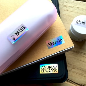 May include: A light pink hard-shell case, a tan notebook, and a black folder, each featuring rectangular holographic name labels. The labels display the names Marvin and Andrew Edwards in black text, with a small lightbulb icon and a smiley face icon.