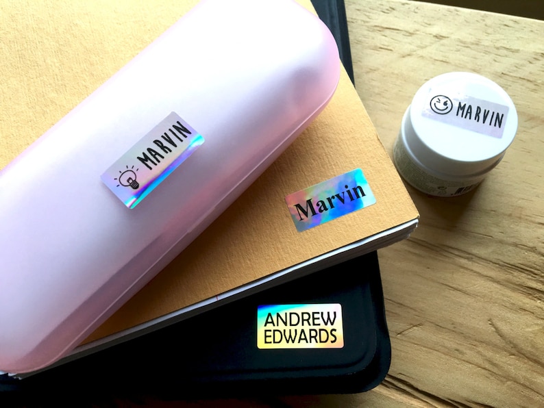 May include: A light pink hard-shell case, a tan notebook, and a black folder, each featuring rectangular holographic name labels. The labels display the names Marvin and Andrew Edwards in black text, with a small lightbulb icon and a smiley face icon.
