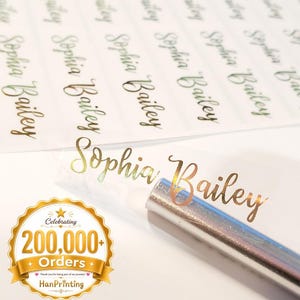 Könnte beinhalten: Ein Bogen mit rechteckigen Klebeetiketten, auf denen der Name Sophia Bailey in einer metallisch goldenen Schreibschrift gedruckt ist. Ein einzelnes Etikett mit demselben Namen befindet sich auf einer silbernen Tube neben einem goldenen Abzeichen mit der Aufschrift Celebrating 200,000+ Orders.