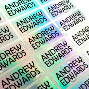 May include: A sheet of rectangular adhesive labels featuring a holographic finish that shifts between green, blue, and silver tones. Each individual label is printed with the name Andrew Edwards in a bold, black, sans-serif capital font.