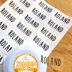 May include: A sheet of rectangular clear adhesive labels featuring the name ROLAND printed in a bold black sans-serif font. A white circular object with a gold and white sticker reading Celebrating 200,000+ Orders HanPrinting is present.