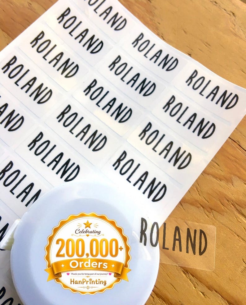 May include: A sheet of rectangular clear adhesive labels featuring the name ROLAND printed in a bold black sans-serif font. A white circular object with a gold and white sticker reading Celebrating 200,000+ Orders HanPrinting is present.