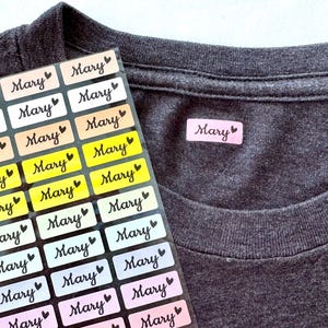 May include: A collection of name labels printed with the name "Mary" in a handwritten style, each with a heart symbol. The labels are in pastel shades. A grey t-shirt has a single label attached.