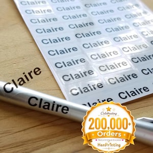 以下が含まれることがあります： 黒でClaireと印刷されたシルバーのペンと、同じくClaireと黒で印刷された長方形の粘着ラベルシート。Celebrating 200,000+ Orders HanPrintingというテキストが書かれた金色の円形のバッジが写っています。