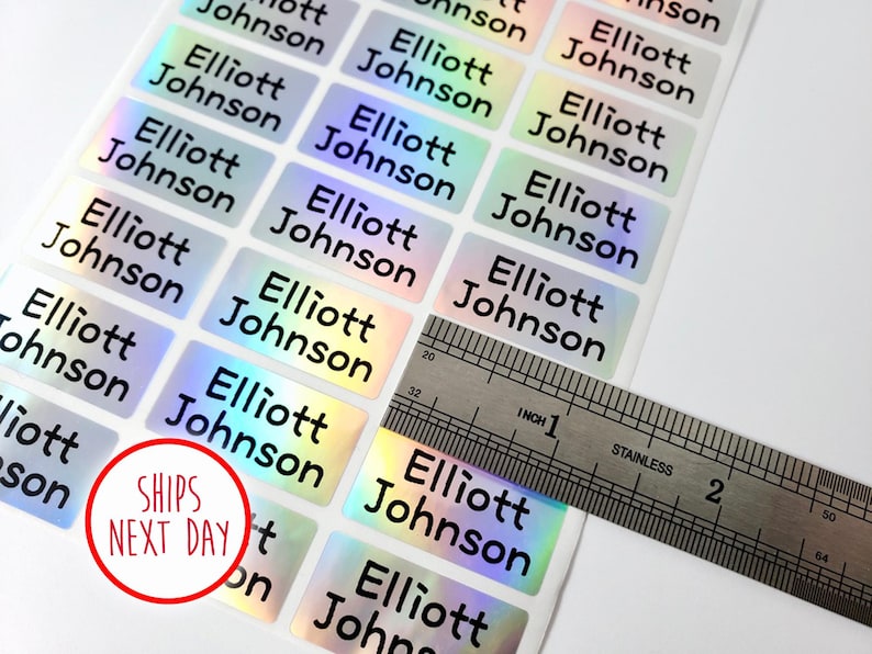 May include: A sheet of rectangular adhesive labels with a holographic finish, each printed with the name Elliott Johnson in black sans-serif font. A stainless steel ruler is placed over the labels, showing a width of approximately 1.5 inches.