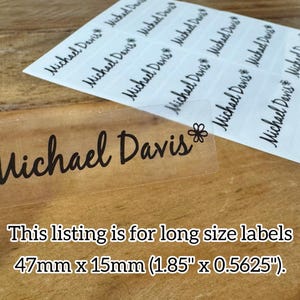 May include: Clear label with the name "Michael Davis" in black script, with a small flower graphic. The label is 1.85" x 0.5625". Several similar labels are visible on a white sheet. The background is a wooden surface.