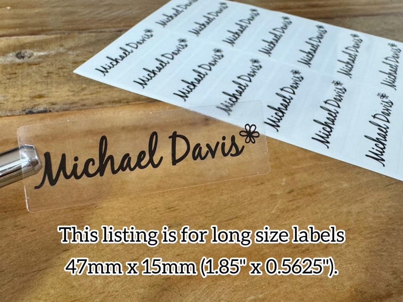 May include: Clear label with the name "Michael Davis" in black script, with a small flower graphic. The label is 1.85" x 0.5625". Several similar labels are visible on a white sheet. The background is a wooden surface.