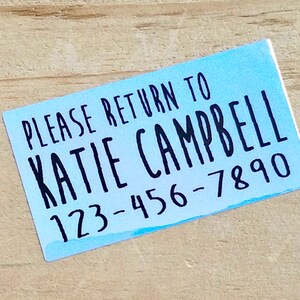 K&ouml;nnte beinhalten: Ein rechteckiger Aufkleber mit hellblauem Aquarellhintergrund. Der schwarze Text lautet "PLEASE RETURN TO KATIE CAMPBELL 123-456-7890". Der Aufkleber befindet sich auf einer hellbraunen Holzoberfl&auml;che.