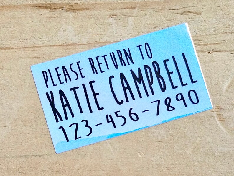 K&ouml;nnte beinhalten: Ein rechteckiger Aufkleber mit hellblauem Aquarellhintergrund. Der schwarze Text lautet "PLEASE RETURN TO KATIE CAMPBELL 123-456-7890". Der Aufkleber befindet sich auf einer hellbraunen Holzoberfl&auml;che.