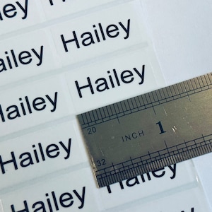May include: A sheet of rectangular adhesive labels featuring the name Hailey printed in a dark blue sans-serif font. A metal ruler is positioned over the labels, showing that each individual label measures approximately 1.5 inches in width.