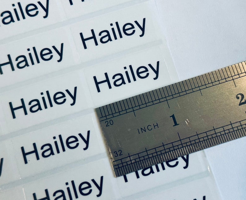 May include: A sheet of rectangular adhesive labels featuring the name Hailey printed in a dark blue sans-serif font. A metal ruler is positioned over the labels, showing that each individual label measures approximately 1.5 inches in width.