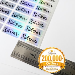 May include: A sheet of rectangular stickers with a holographic finish, each featuring the word Silver in a black cursive script. A metal ruler is positioned across the bottom, showing the stickers are approximately 1 inch in width.