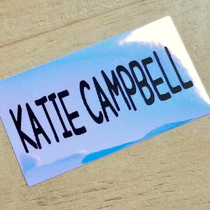 K&ouml;nnte beinhalten: Ein rechteckiger Aufkleber mit einem hellblauen und wei&szlig;en Farbverlauf. Der Aufkleber tr&auml;gt den Namen "KATIE CAMPBELL" in fetter, schwarzer, handschriftlicher Schrift.