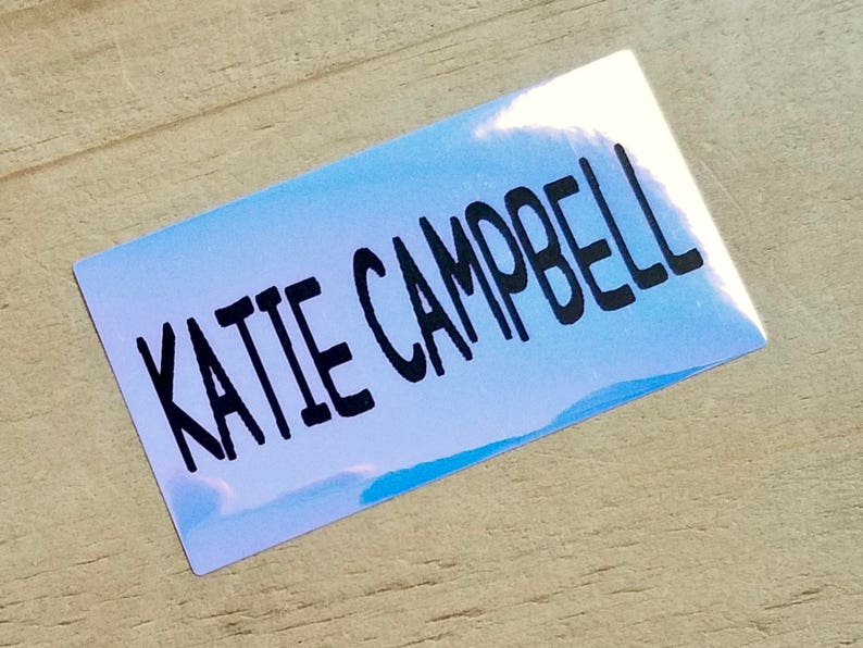 K&ouml;nnte beinhalten: Ein rechteckiger Aufkleber mit einem hellblauen und wei&szlig;en Farbverlauf. Der Aufkleber tr&auml;gt den Namen "KATIE CAMPBELL" in fetter, schwarzer, handschriftlicher Schrift.