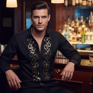 Puede incluir: Una camisa negra con bordados dorados, usada por una persona en un bar. Un vaso de líquido ámbar está en la barra. Un camarero está en el fondo.