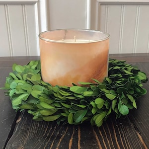 May include: A large, amber-coloured candle with a white wax top sits in a green boxwood wreath on a dark wood surface.