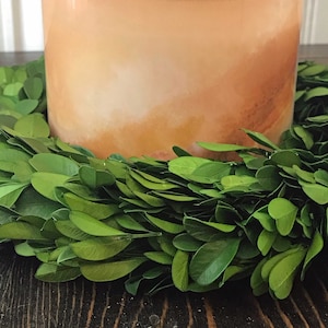 May include: A clear glass candle jar with a light orange and brown ombre design. The candle is surrounded by a green boxwood wreath.