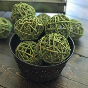 May include: A black metal bowl filled with green moss balls. The bowl has a distressed finish and is sitting on a dark wood surface.