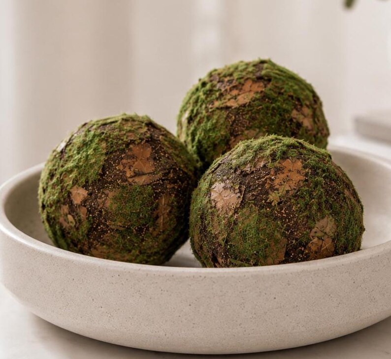 May include: Three spherical decorative balls covered in green moss and brown textured material, arranged inside a shallow, round, off-white ceramic bowl with a speckled finish. The mossy spheres have a natural, uneven surface texture.