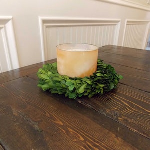 May include: A brown wooden table with a green boxwood wreath and a clear glass candle holder with a white candle inside.