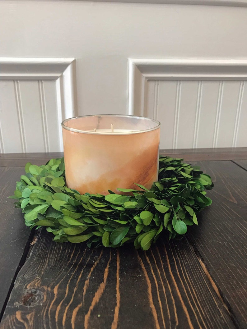 May include: A large, amber-coloured candle with a white wax top sits in a green boxwood wreath on a dark wood surface.