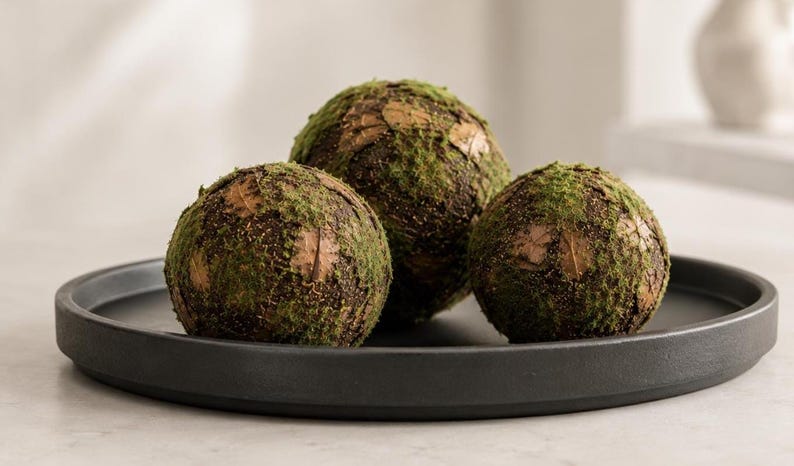 May include: Three spherical decorative objects covered in a textured layer of green moss and brown bark-like material. The spheres are arranged together on a flat, circular, matte black tray with a raised edge, creating a cohesive display.