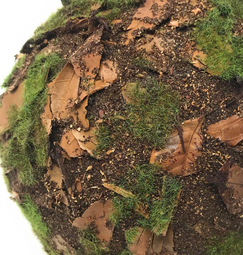 May include: A spherical decorative object covered in a textured layer of green artificial moss, scattered brown dried leaves, and dark brown soil-like particles, creating a dense, forest-floor appearance across the entire rounded surface.
