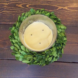 May include: A three-wick candle in a clear glass jar with yellow wax and three wicks. The candle is sitting on a green boxwood wreath.