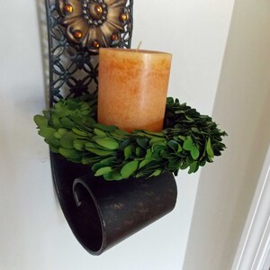 May include: A black metal wall sconce with an ornate design holds a candle and a green boxwood wreath.