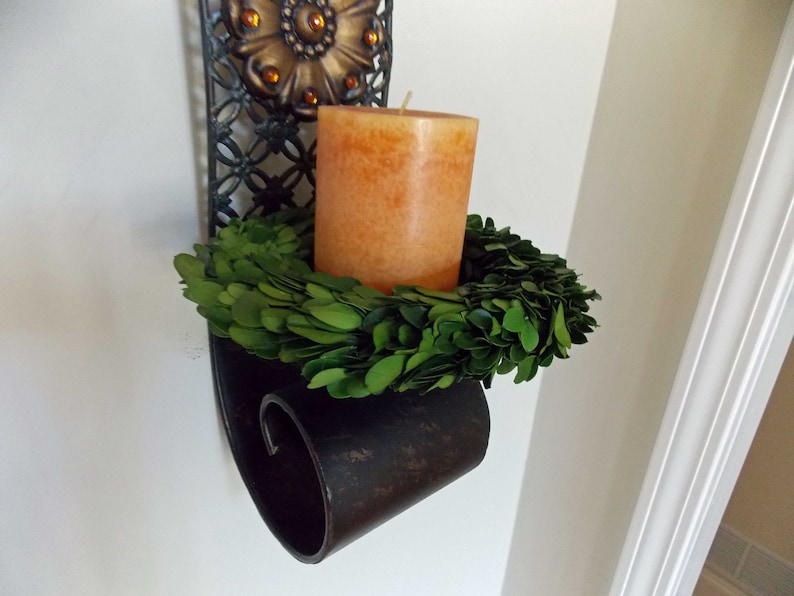 May include: A black metal wall sconce with an ornate design holds a candle and a green boxwood wreath.