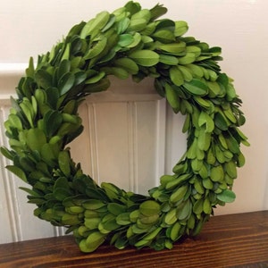 May include: A green boxwood wreath with lush, full foliage. The wreath is made of natural boxwood branches and is perfect for adding a touch of greenery to any space.