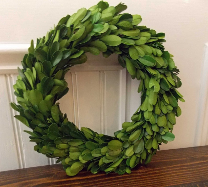 May include: A green boxwood wreath with lush, full foliage. The wreath is made of natural boxwood branches and is perfect for adding a touch of greenery to any space.