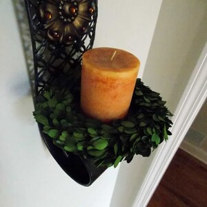 May include: A black metal wall sconce with a decorative floral design holds a round, orange candle surrounded by a green boxwood wreath.