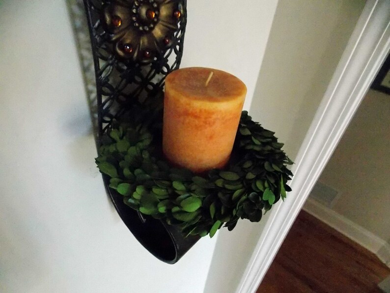 May include: A black metal wall sconce with a decorative floral design holds a round, orange candle surrounded by a green boxwood wreath.