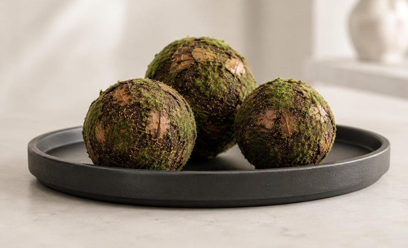 May include: Three spherical decorative objects covered in green moss and brown textured material, arranged together on a circular matte black tray with a raised edge. The spheres feature a mix of dark brown patches and vibrant green moss.