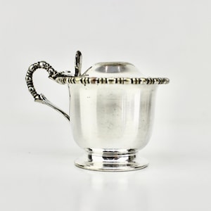 May include: A small, silver-coloured creamer with a lid and a handle. The creamer has a decorative beaded rim and a small spoon resting on the lid. The handle is ornate, and the creamer sits on a small, round base. The background is white.