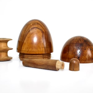 May include: A wooden puzzle with six pieces, including a dome, a base, a cylinder, and three smaller pieces. The pieces are made of light brown wood with a smooth finish.