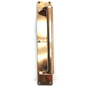 Könnte beinhalten: Ein antiker Türgriff aus Messing mit rechteckiger Grundplatte und gebogenem Griff. Das Messing hat eine warme Patina. Der Türgriff ist auf weißem Hintergrund montiert. Ein klassisches Element der Architektur.