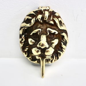 May include: A brass lion head door knocker with a detailed mane and a long, pointed striker. The knocker is a warm gold colour and is mounted on a white surface.