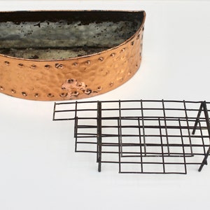 May include: A copper-coloured, half-round planter with a hammered texture and rivet details. A separate, black metal grid is also present, likely for plant support. The items are on a white background.