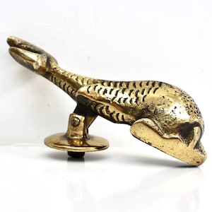 May include: A brass door knocker, shaped like a stylized animal, possibly a hare or rabbit, with etched details. The knocker is mounted on a round base. The background is white.