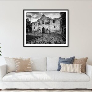 May include: A black and white framed wall art print depicting the Alamo mission building with its stone facade and arched entrance, set against a cloudy sky, displayed above a white sofa with various decorative throw pillows in a living room.