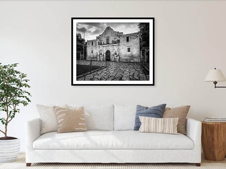 May include: A black and white framed wall art print depicting the Alamo mission building with its stone facade and arched entrance, set against a cloudy sky, displayed above a white sofa with various decorative throw pillows in a living room.