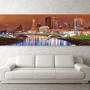 May include: A panoramic view of a city skyline at night with a bridge in the foreground. The city is lit up with colorful lights, and the bridge is reflected in the water. The buildings are tall and modern, and the sky is a deep orange.