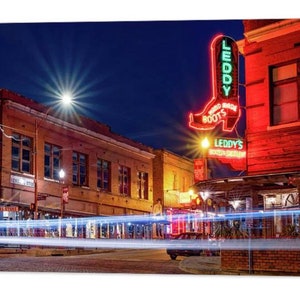 May include: A night scene of a city street with a neon sign for "Leddy's Boot Shop" in the shape of a red cowboy boot. The sign reads "Hand Made Boots". The street is lit with streetlights and car headlights.