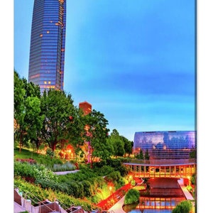 May include: A view of a modern city skyline at dusk with a tall glass building and a large glass structure with a reflecting pool in the foreground. The pool is surrounded by lush green landscaping and a walkway.