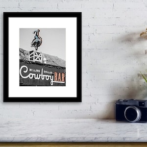 May include: Black and white photo of a cowboy on a bucking bronco atop a building with a sign that reads "Million Dollar Cowboy Bar".