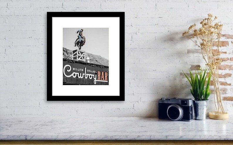 May include: Black and white photo of a cowboy on a bucking bronco atop a building with a sign that reads "Million Dollar Cowboy Bar".